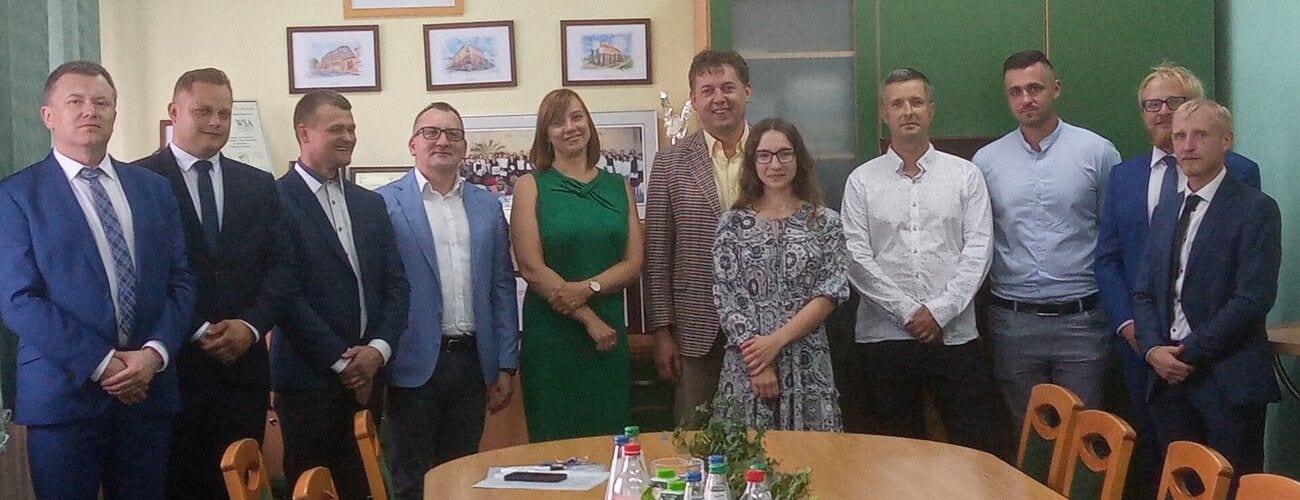Absolwenci i przedstawiciele uczelni pozujący w sali przy stole egzaminacyjnym.