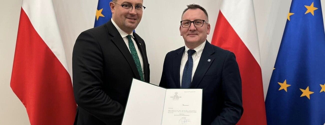 Dwaj mężczyźni prezentujący dokument nominacji na tle flag Polski i Unii Europejskiej.