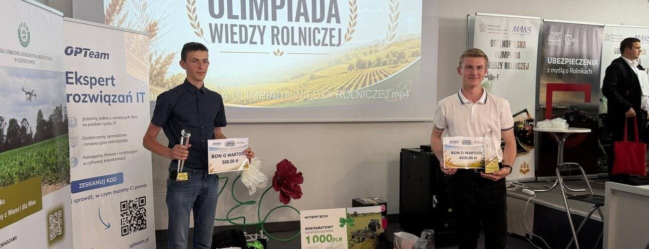 Dwaj młodzi mężczyźni z dyplomami i nagrodami na tle planszy Olimpiady Wiedzy Rolniczej.