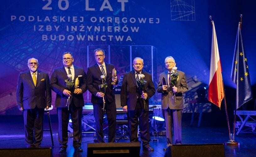 Uczestnicy gali 20-lecia Podlaskiej Okręgowej Izby Inżynierów Budownictwa na scenie z kwiatami i wyróżnieniami.