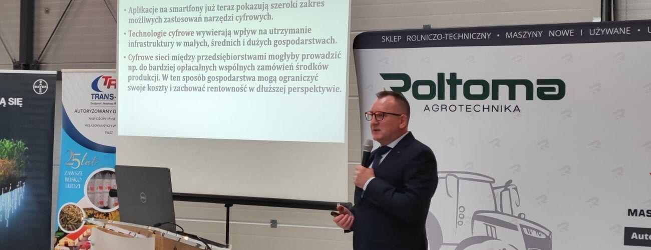 Prelegent z mikrofonem na tle slajdu prezentacji i planszy firmy rolniczej.