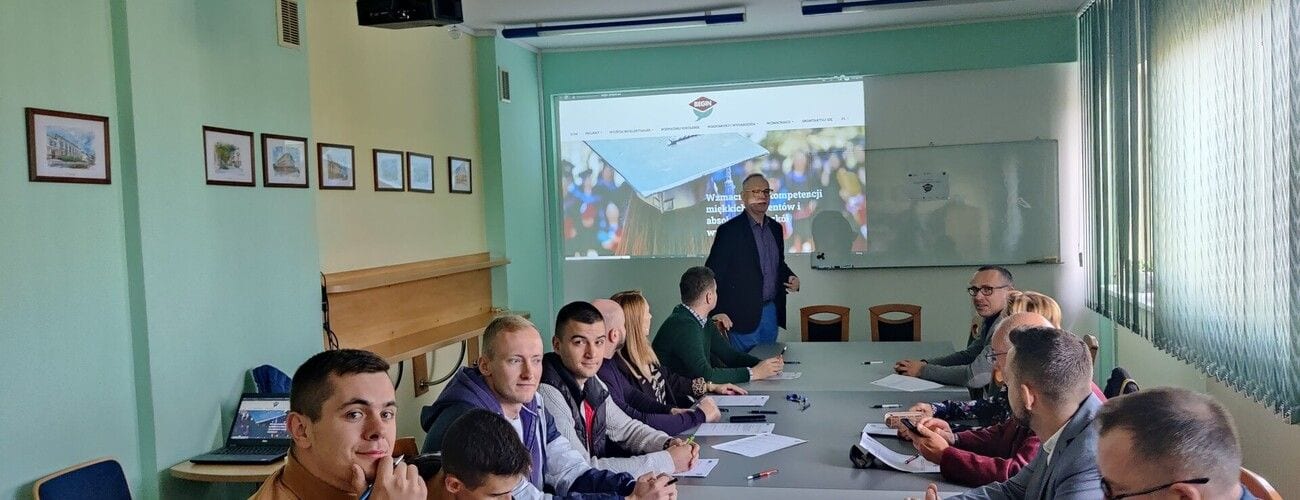 Uczestnicy spotkania projektu BEGIN przy stołach w sali z ekranem prezentacyjnym.