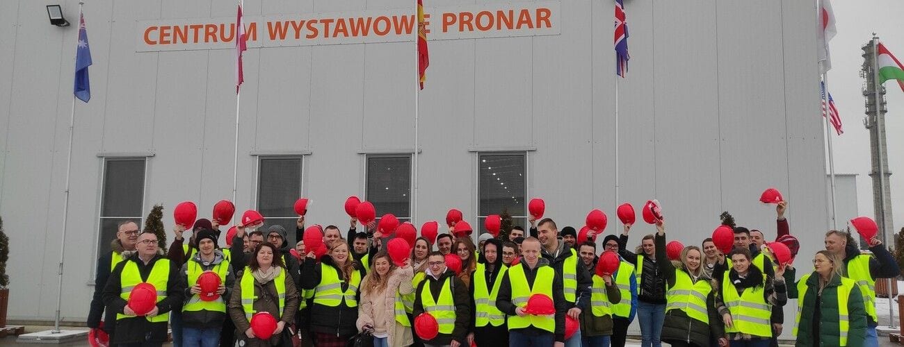Grupa w kamizelkach odblaskowych unosi czerwone kaski przed Centrum Wystawowym Pronar.