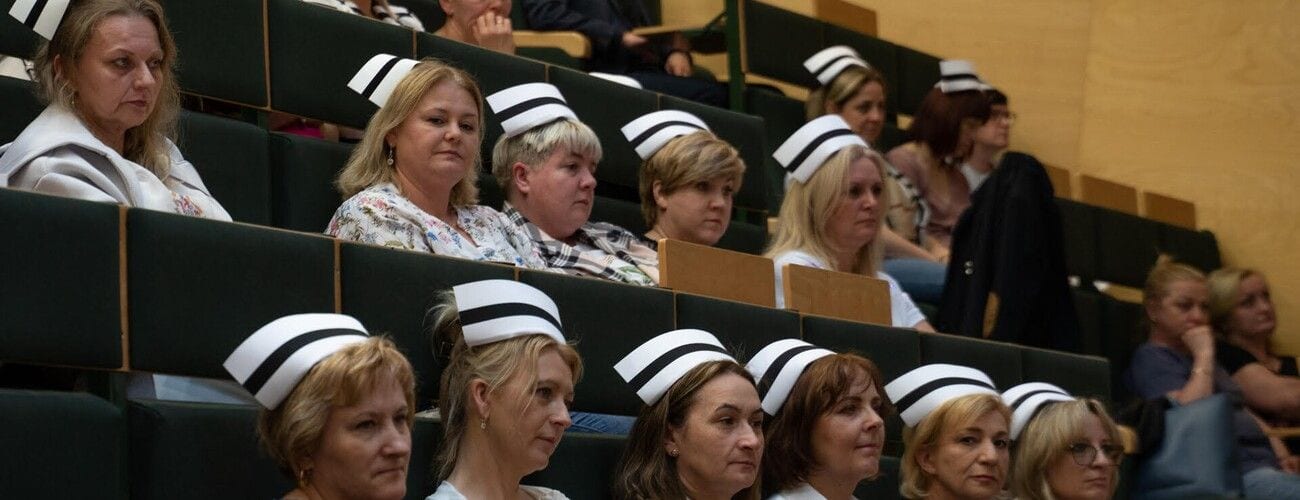 Uczestniczki uroczystości pielęgniarskiej siedzące w auli, część w białych czepkach z czarnym paskiem.