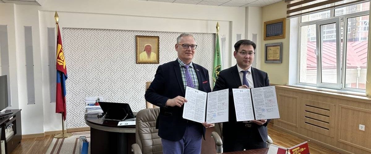 Dwaj przedstawiciele instytucji prezentujący otwarte dokumenty porozumienia, na tle flag.