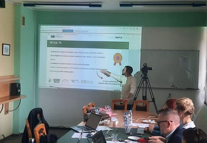 Prelegentka wskazuje slajd podczas spotkania projektu KICRO; uczestnicy siedzą przy stole.
