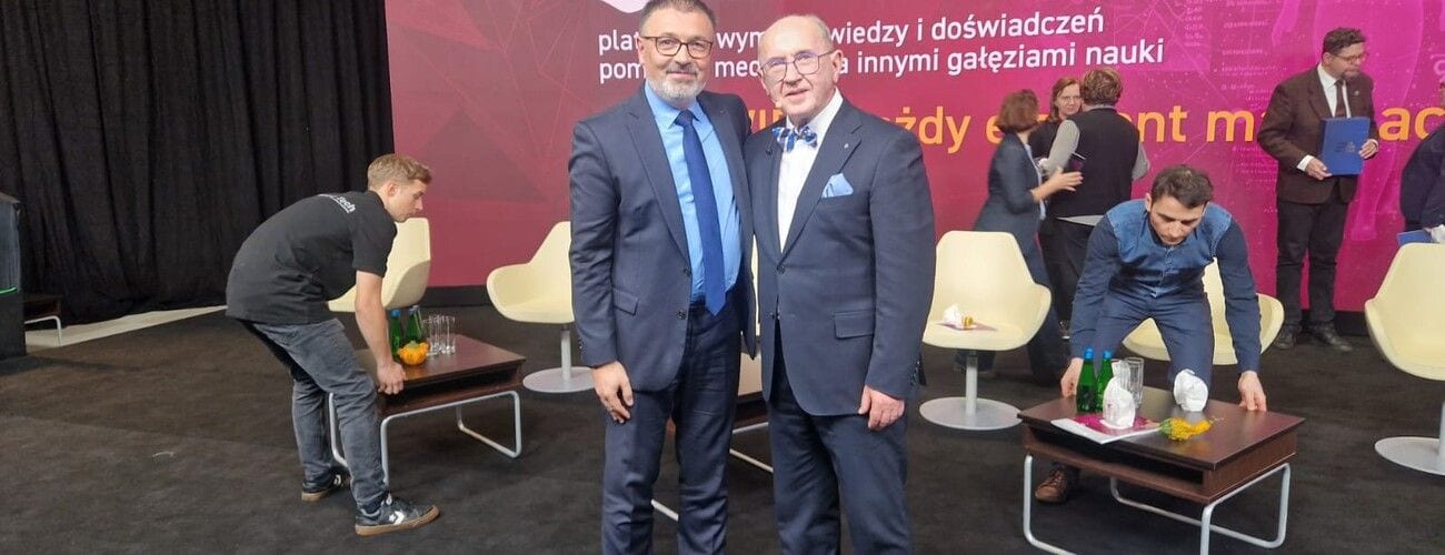 Dwaj mężczyźni w garniturach pozujący na scenie konferencyjnej.