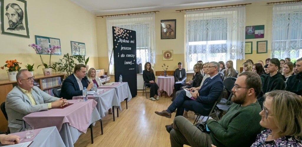 Uczestnicy spotkania literackiego w szkolnej sali z portretem Norwida i dekoracjami.