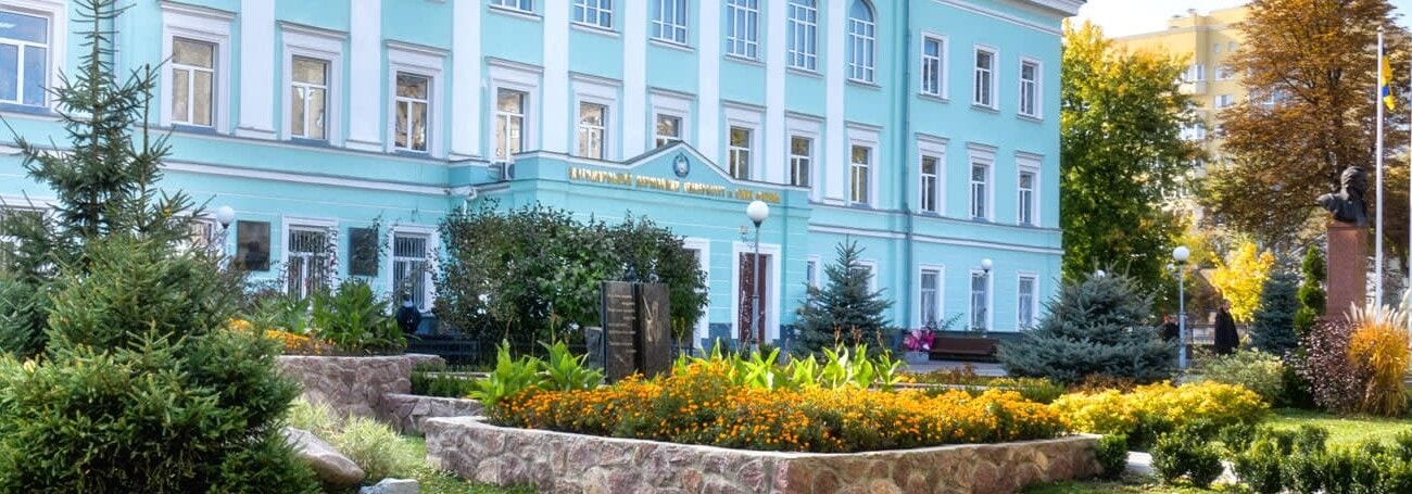 Jasnoniebieski budynek uczelni otoczony drzewami i ozdobną zielenią.