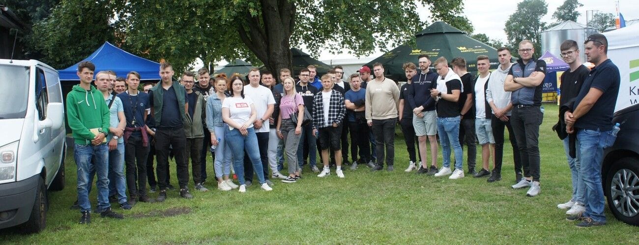 Grupa uczestników zajęć terenowych na trawniku między samochodami i namiotami wystawowymi.