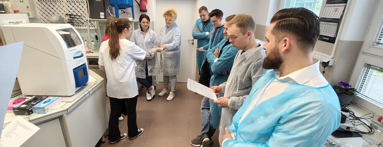 Uczestnicy wizyty w laboratorium w białych i jasnoniebieskich fartuchach, przy aparaturze badawczej.