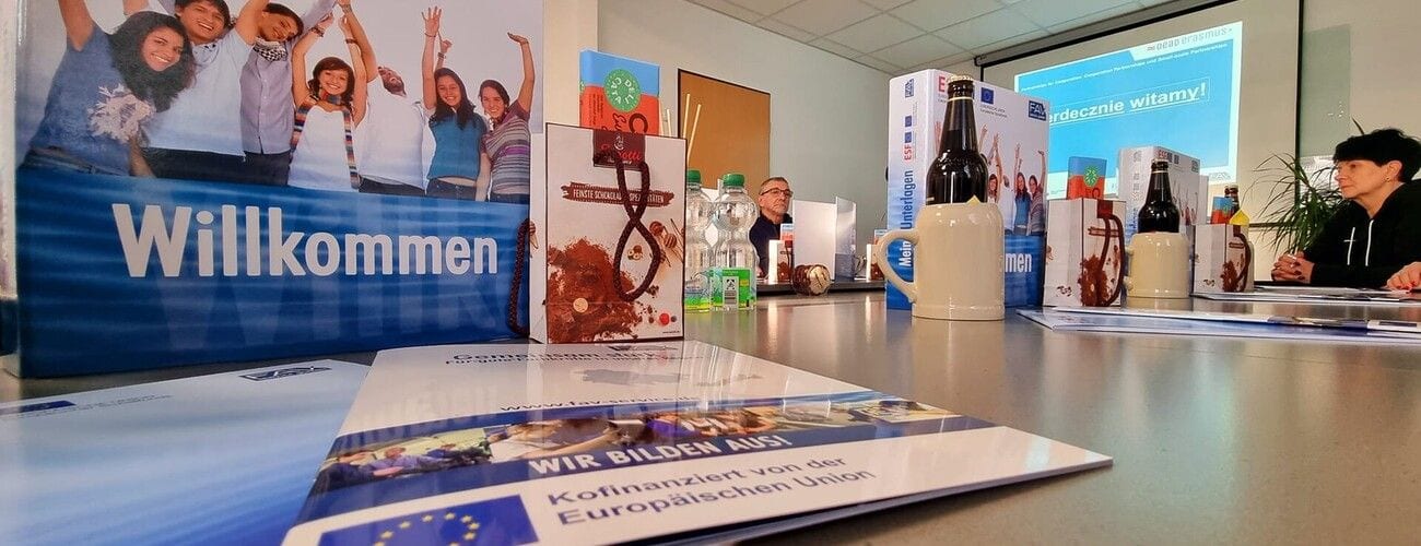 Sala szkoleniowa z napisem „Willkommen”, materiałami i uczestnikami przy stole.