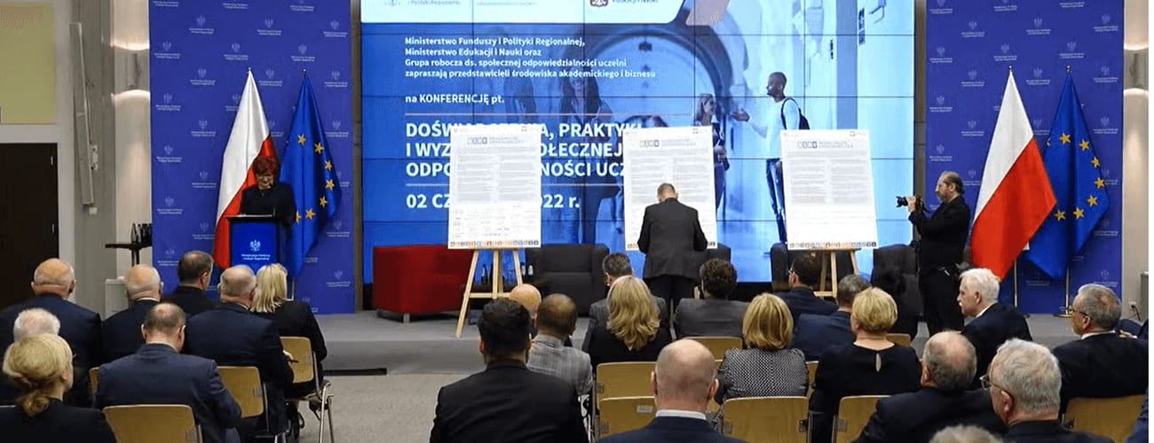 Uroczystość z dokumentami Deklaracji Społecznej Odpowiedzialności Uczelni na scenie oraz publicznością na sali.