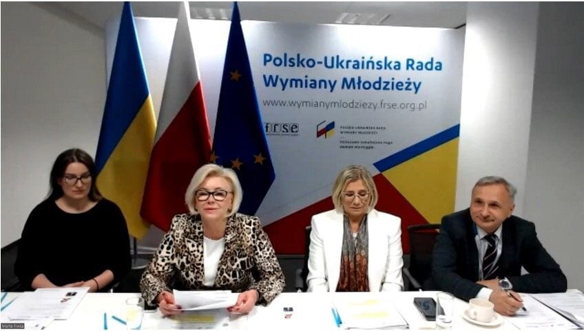 Cztery osoby przy stole na tle flag i planszy Polsko-Ukraińskiej Rady Wymiany Młodzieży.