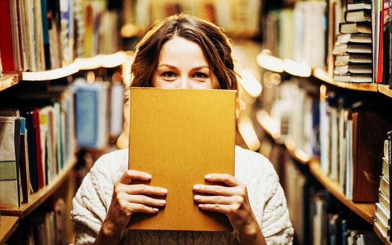 Kobieta trzymająca otwartą książkę przed twarzą między regałami bibliotecznymi.
