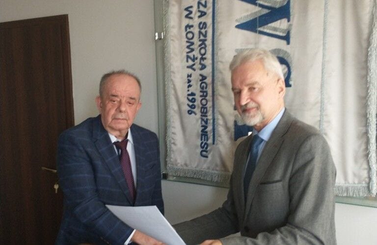 Dwaj przedstawiciele instytucji z dokumentem porozumienia na tle sztandaru uczelni.