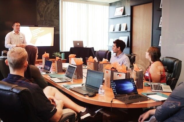 Prelegent i uczestnicy międzynarodowego spotkania przy stole z laptopami.