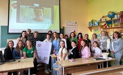 Uczestnicy warsztatów edukacyjnych pozujący w sali z pomocami dydaktycznymi i ekranem wideokonferencji.