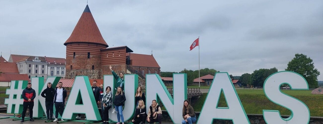 Grupa pozująca przy dużym napisie „Kaunas” na tle zamku w Kownie.