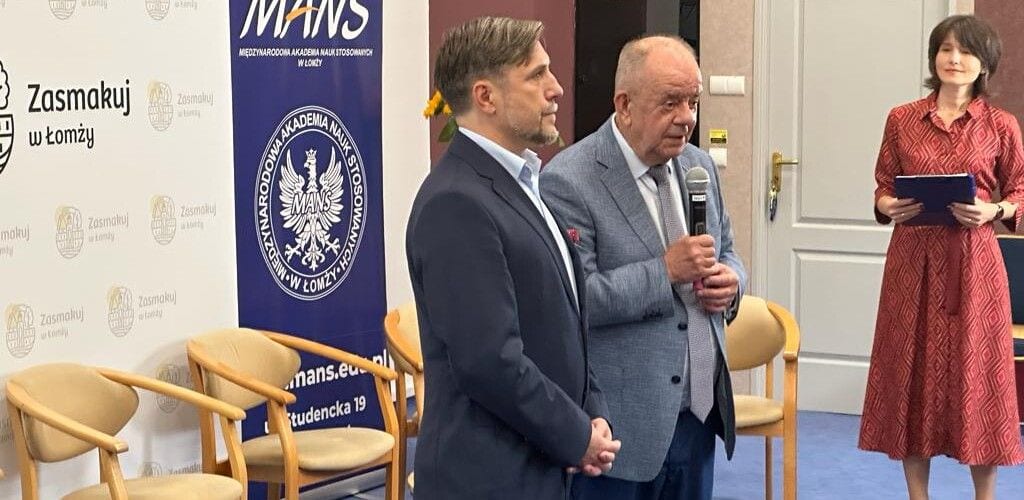 Prowadzący spotkanie „Debatowanie na śniadanie” przy mikrofonie na tle plansz MANS i miasta Łomża.