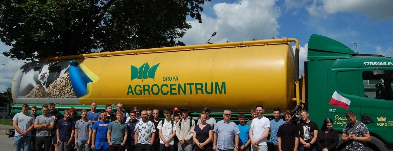 Studenci na tle żółtego pojazdu do transportu pasz z oznaczeniem Agrocentrum.