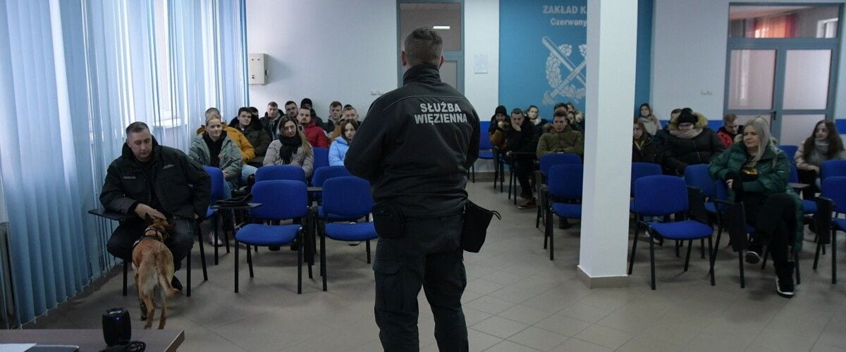 Funkcjonariusz Służby Więziennej prowadzi spotkanie ze studentami; obok znajduje się przewodnik z psem.