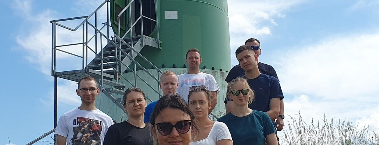 Uczestnicy zajęć terenowych przy zielonej instalacji przemysłowej z metalowymi schodami.