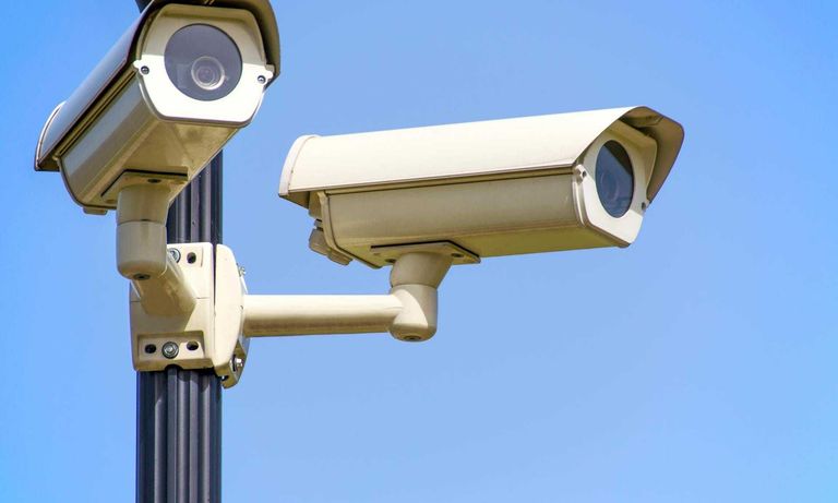 Two beige security cameras are mounted on a black pole against a bright blue sky.