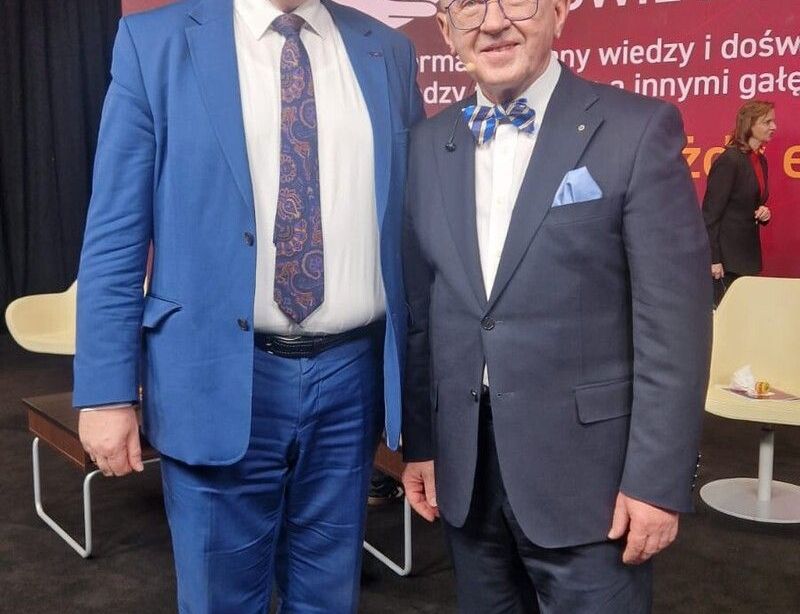 Two smiling men in suits stand together at a conference, looking at the camera.
