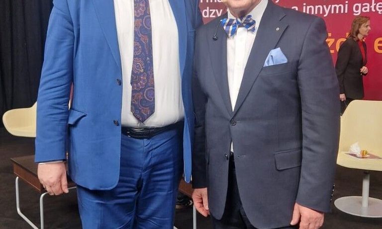 Two smiling men in suits stand together at a conference, looking at the camera.